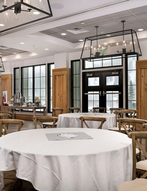 A bright event hall with round tables, wooden chairs, and hanging lantern-style lights, set up for a dining or banquet.