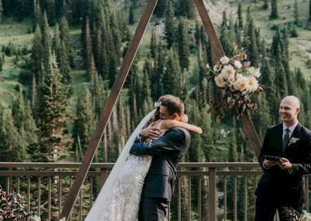 An outdoor wedding: a bride and groom embrace under a floral arch as a suited man stands by in a mountain valley.