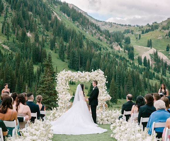 A wedding ceremony with a bride and groom at an outdoor arch of white flowers, surrounded by guests in a mountain valley.