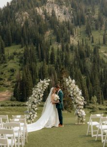 A wedding couple in front of tall pine-covered mountains, surrounded by white floral arrangements and chairs on a grassy outdoor lawn.